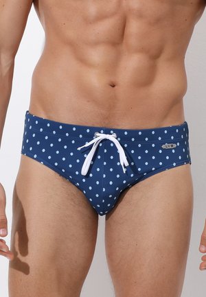 Torace maschile che indossa slip da bagno blu con pois bianchi e coulisse bianca annodata davanti.