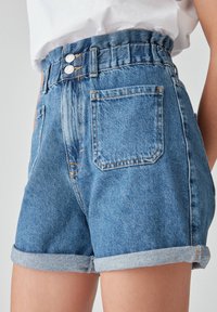 Bermudas de mezclilla de tiro alto con cierre de doble botón, combinadas con una camisa blanca metida por dentro, mostrando los bolsillos frontales y laterales.
