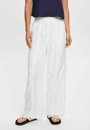 Osoba nosząca białe spodnie z szerokimi nogawkami, czarne i beżowe sandały oraz granatowy krótki top na jednolitym jasnym tle.