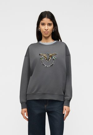 Nő sötétszürke pulóvert visel, amelyen flitteres madármotívum látható, és sötétkék farmert, miközben egy sima, világos háttér előtt áll.