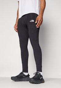 Svarta träningsleggings med en åtsittande design, prydda med en liten vit logotyp på höften, i kombination med svarta löparskor och vita strumpor.