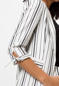 Blazer a righe bianche e nere con maniche a tre quarti, caratterizzato da un dettaglio annodato sui polsini e un design dal fit rilassato.