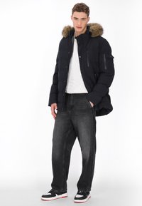 Marineblå pufferjakke med hætte foret med imiteret pels, udstyret med lommer med lynlås og knaplukning; kombineret med grå jeans og sorte sneakers.