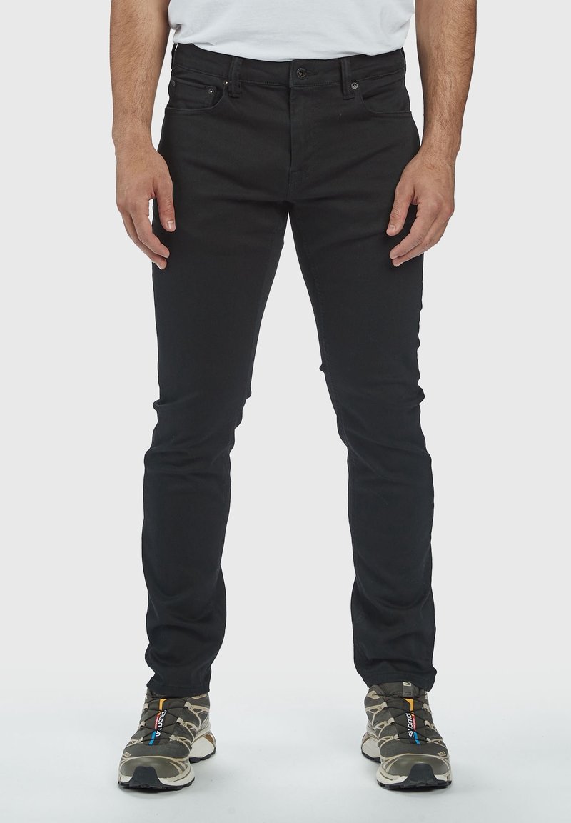 Jeans neri aderenti in denim, con cinque tasche, passanti per cintura e chiusura con zip. Indossati con sneakers a fantasia.