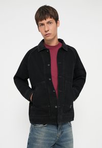 Black denim jacket with a button front, two front pockets, and a collared neck. Worn over a maroon t-shirt and paired with blue jeans.
