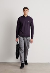 Dunkellila Pullover, graue Hose, schwarze Schuhe. Das Modell hält eine schwarze Tasche, und ein Kragen ist unter dem Pullover sichtbar. Minimalistisches Design.