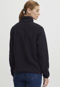 Sort fleece sweater med høj krave, blød tekstur og lange ærmer. Har en let nedfaldende kant og et lille logo-detail på ryggen.