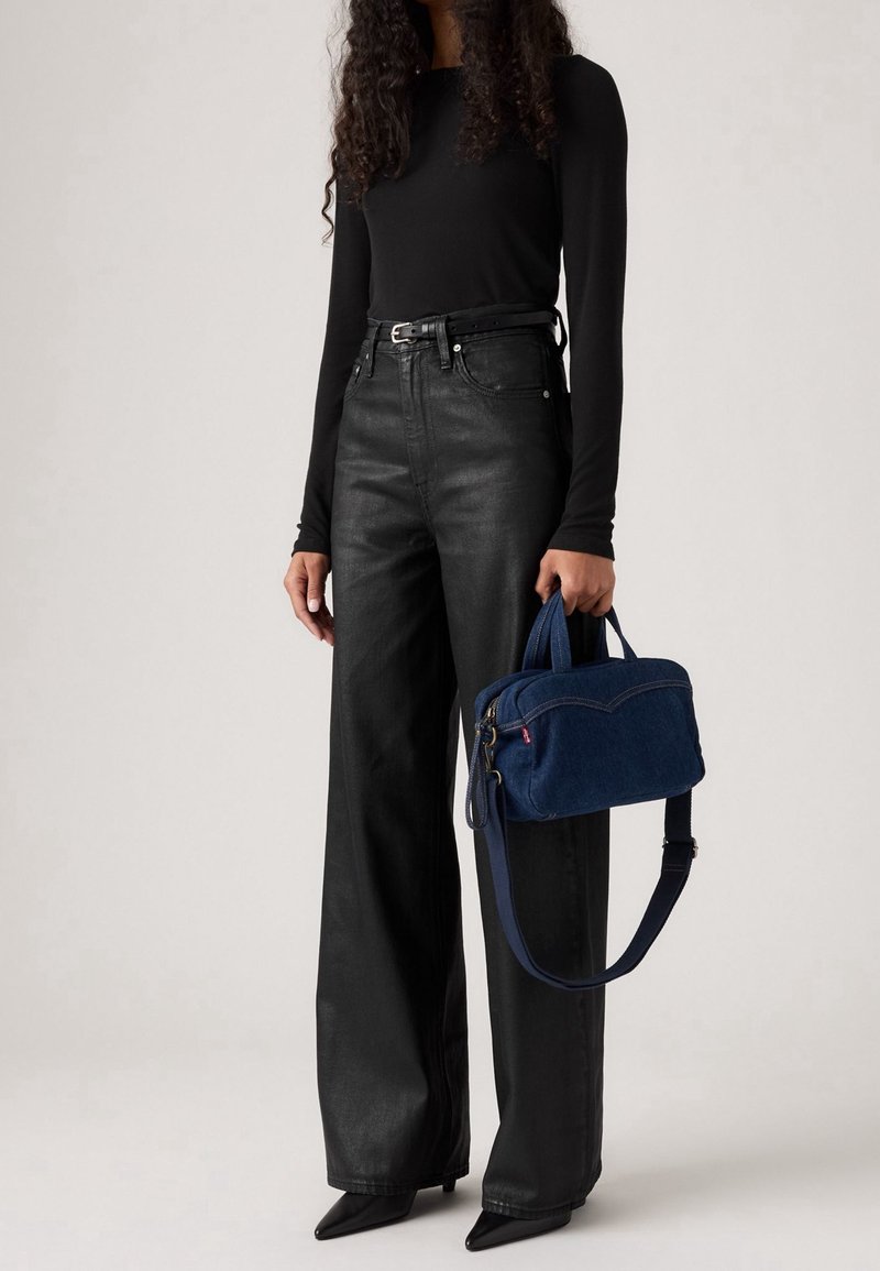 Femme portant un haut noir à manches longues et un pantalon noir taille haute à jambes larges, tenant un sac à main bleu avec des poignées courtes et une bandoulière.