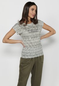 Camiseta de manga corta y ajustada en gris claro con un patrón geométrico. Texto en negro que dice "Disfruta de las pequeñas cosas". Combinada con pantalones verde oliva.