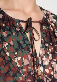 Top blouson multicolore con stampa floreale nei toni del verde, marrone e crema. Presenta una scollatura a occhiello con un nodo e spalle arricciate.