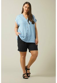 Camisa de botones azul claro con mangas cortas enrolladas, combinada con pantalones cortos negros. Sandalias negras con hebillas, de corte relajado y diseño casual.