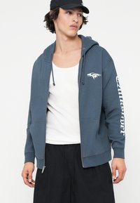 Plava zip-up dukserica od mješavine pamuka, s bijelim logotipom na lijevoj strani grudi i dizajnom teksta na desnoj rukavu. Glatka tekstura, opušteni kroj.