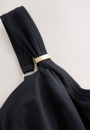 Musta kangasranneke, jossa solmittu solmu ja kullanvärinen metallikoriste. Rannekkeessa on sileä pinta ja hienovaraiset ompeleet.