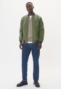Olive green bomber jacket with zip closure, ribbed cuffs, worn over a beige top and blue jeans. Model stands against a white background.