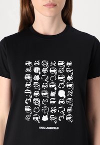 Juodas medvilnės marškinėliai su balta spauda, vaizduojančia įvairių kaukės panašių veidų tinklelį, o apačioje yra tekstas "KARL LAGERFELD".