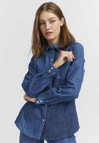 Camicia in denim di blu medio con colletto classico, bottoni frontali e maniche lunghe con polsini abbottonati. Tessuto testurizzato con vestibilità rilassata.