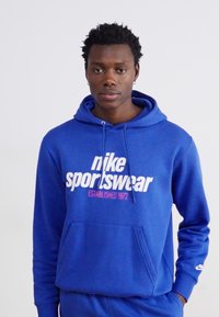 Felpa blu in cotone con tasca frontale, cappuccio con cordino e testo bianco "nike sportswear" con accenti rossi. Logo Nike sulla manica.
