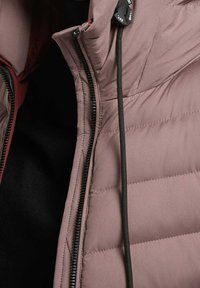 Gros plan d'une veste matelassée mauve avec une fermeture éclair noire partiellement ouverte et un cordon noir avec un toggles de marque au col.