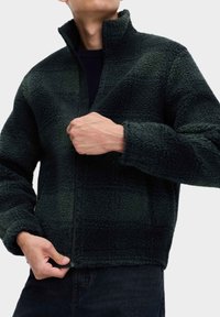 Polarowa kurtka zapinana na zamek w ciemnozielonym kolorze, z wysokim kołnierzem, teksturowaną powierzchnią i elastycznym dołem. Zamek błyskawiczny z przodu.