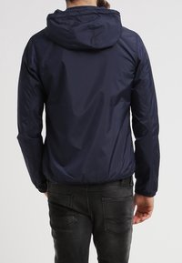 Marinblå lätt jacka med huva, slät textur, elastiska ärmslut och en något rynkad nederkant, bärs över mörka jeans.