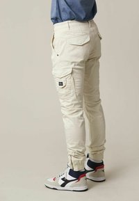 Pantalons cargo beige clair avec des poignets élastiques, plusieurs poches et une petite étiquette de marque sur le côté, associés à des baskets montantes blanches et marine.
