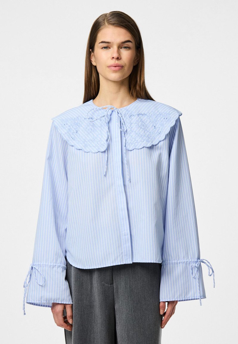 Ανοιχτό μπλε ριγέ τοπ με μεγάλο κεντητό γιακά, μακριά μανίκια με κλείσιμο δέσιμο και χαλαρή εφαρμογή. Λείο υφασματικό υλικό.