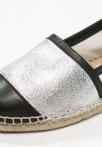 Espadrille noire et argentée avec un dessus métallique texturé, des accents en cuir noir et une semelle en jute avec des coutures décoratives.