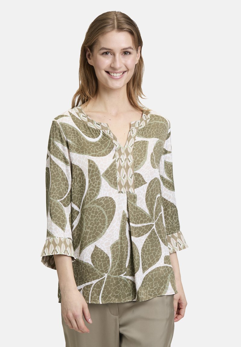 Jonge vrouw die glimlacht, in een losse groene en witte geblokte blouse met 3/4 mouwen en beige broek, staande tegen een neutrale achtergrond.
