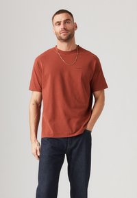 Camiseta de manga corta en color óxido, hecha de algodón, con un cuello redondo y un logo sutil en el pecho. Combinada con jeans oscuros.
