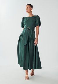 Pitkä vihreä mekko, jossa on puhvihihat, vyötärönauha, jossa on metallinen solki, koottu hame ja sileä pinta. Valkoiset hihnasandaalit.