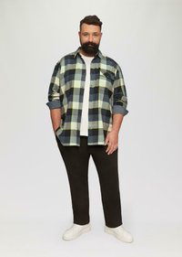 Geruit flanellen overhemd in groen, zwart en geel over een wit t-shirt, gecombineerd met zwarte broek en witte sneakers. Modern, casual fit.