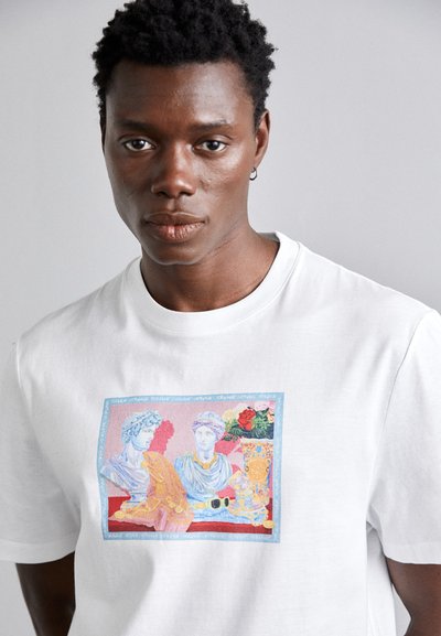 Camiseta blanca de algodón con un estampado gráfico colorido que presenta esculturas clásicas y acentos florales, todo ello sobre un fondo en contraste.