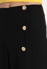 Jupe taille haute noire avec trois grands boutons dorés à l'avant, associée à un haut jaune visible à la taille.