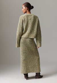 Pleteni komplet u prigušenoj zelenoj boji, s kraćim džemperom s rebrastim rubom i dugom, pripijenom suknjom. Upotpunjeno šiljastim smeđim čizmama.