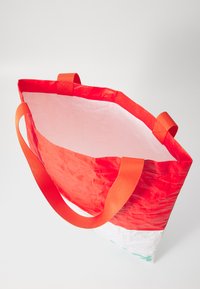 Sac fourre-tout réutilisable rouge et blanc en matériau texturé et durable, doté de deux poignées orange pour le transport. Ouvert en haut, sans fermeture.