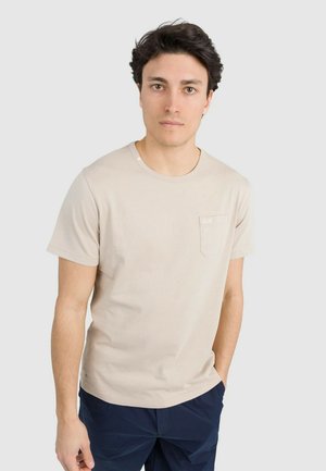 Giovane uomo che indossa una T-shirt beige a maniche corte con taschino sul petto e pantaloni blu navy, in piedi contro uno sfondo chiaro e uniforme.