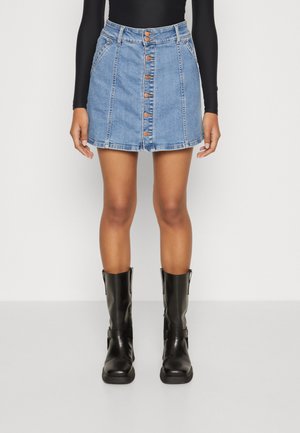 Donna che indossa una gonna di jeans con bottoni davanti, una maglietta nera a maniche lunghe e stivali in pelle neri fino a metà polpaccio, in piedi davanti a uno sfondo bianco.