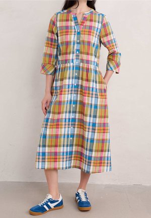 Kobieta ubrana w midi sukienkę w wielokolorową kratę z guzikami, z rękawami do łokcia oraz niebieskie i białe trampki.