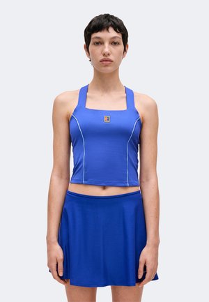 Femme aux cheveux courts et foncés portant un crop top bleu sans manches et une jupe bleue assortie, debout devant un fond clair uni.