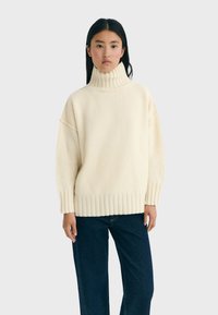 Cremfarbener Rollkragenpullover aus weichem Strickmaterial mit gerippten Bündchen und Saum, kombiniert mit dunkelblauen Jeans. Sichtbare lockere Passform.