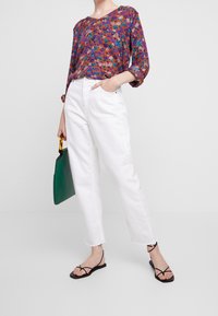 Top a fantasia floreale in viola, arancio, blu e verde, abbinato a jeans bianchi a gamba dritta e sandali neri con cinturini. Borsa verde inclusa.