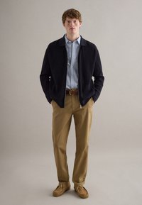 Joven con chaqueta negra con cremallera, camisa de botones azul con rayas, cinturón marrón, pantalones caqui y mocasines beige, de pie con las manos en los bolsillos.