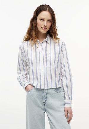 Młoda kobieta o falowanych brązowych włosach, ubrana w białą koszulę na guziki w niebieskie pionowe paski oraz jasnoniebieskie dżinsy, stojąca z jedną ręką w kieszeni.