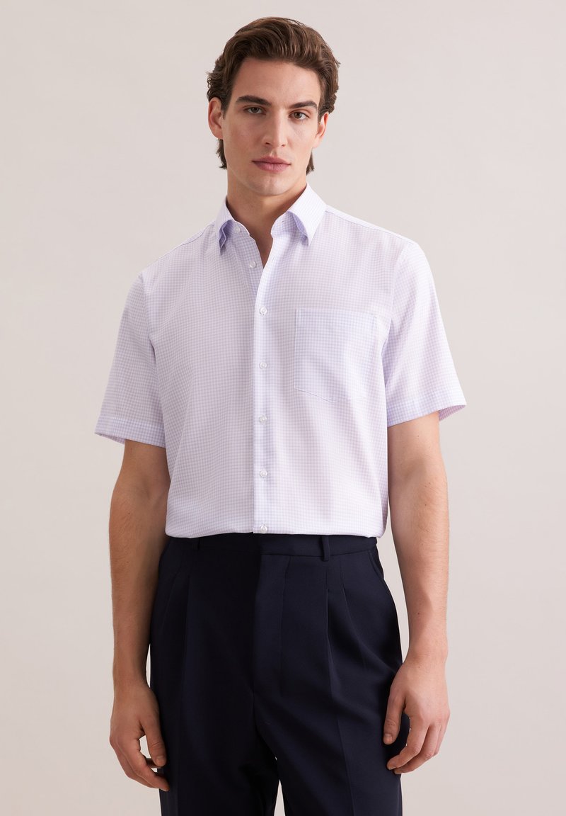 Jeune homme portant une chemise à manches courtes de couleur violet clair avec une poche poitrine et un pantalon plissé foncé, posé devant un fond uni.