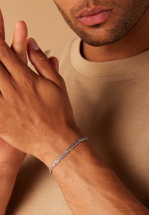 Silver chain bracelet with interlinked oval and flat links, worn on a tanned wrist, against a neutral background.
