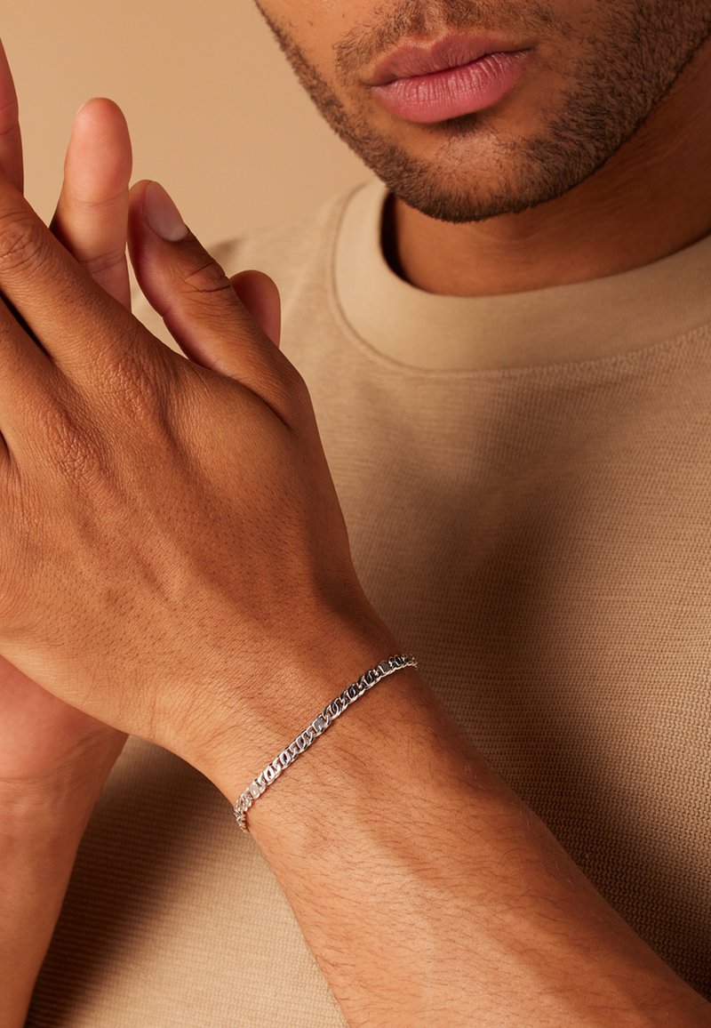Bracelet en chaîne argentée avec des maillons ovales et plats entrelacés, porté sur un poignet bronzé, sur un fond neutre.