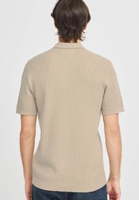 Beige kötött pólóing rövid ujjal, bordázott textúrával és egyenes szegéllyel. Klasszikus galléros kialakítás finom kötött részletekkel.