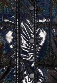 Schwarze Steppjacke mit glänzender, reflektierender Oberfläche und schillernden Sprenkeln in Blau, Grün und Orange. Mit schwarzen Knöpfen und Kordelzug.