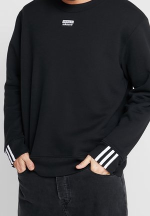 Sort Adidas sweatshirt med hvid logo-patch på brystet og hvide tre-stribe detaljer på ærmekanter, båret med sorte jeans.