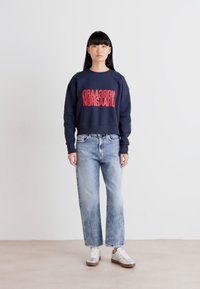 Sudadera navy con texto rojo invertido, combinada con jeans de pierna ancha de lavado claro y zapatillas blancas. Diseño minimalista, texturas suaves.